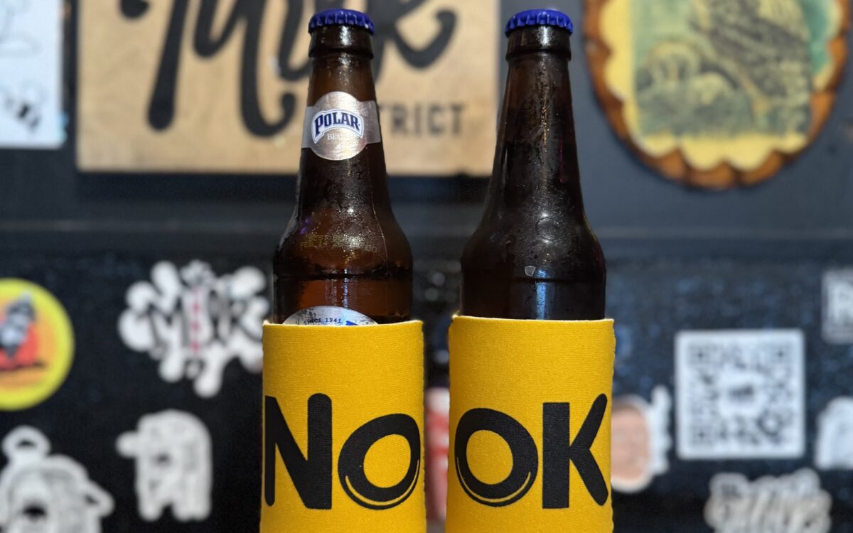 Beer bottles in yellow NOOK koozies with Milk District wall behind