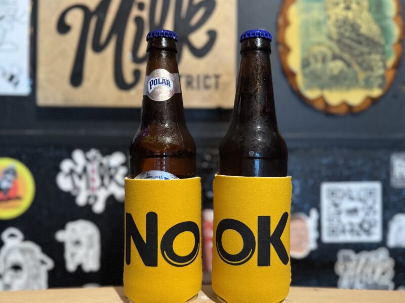 Beer bottles in yellow NOOK koozies with Milk District wall behind